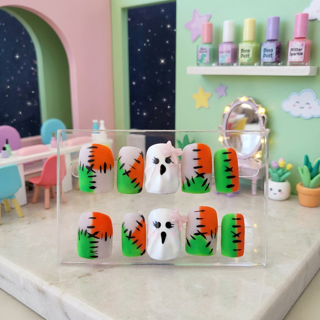 A set of Halloween-themed press-on nails displayed in a pink butterfly-patterned box. The nails feature a mix of orange, green, white, and gray designs, including two ghost designs and striped stitched-look nails.