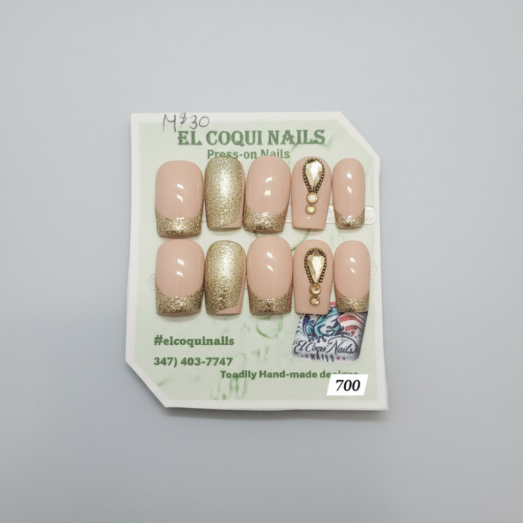 A set of 10 press-on nails displayed on a branded card. The nails feature a nude beige base with metallic gold glitter accents on the tips and sides, plus one nail with a gold teardrop charm design.