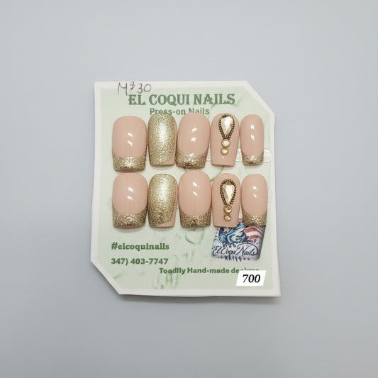 A set of 10 press-on nails displayed on a branded card. The nails feature a nude beige base with metallic gold glitter accents on the tips and sides, plus one nail with a gold teardrop charm design.