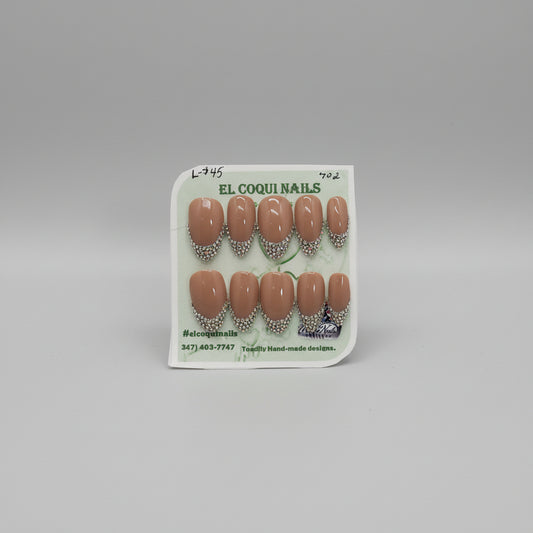 A set of 10 press-on nails on a display card, featuring a glossy nude brown base with a silver rhinestone French tip design. The brand 'EL COQUI NAILS' is printed on the card, and a white label at the bottom displays the style number '702'.