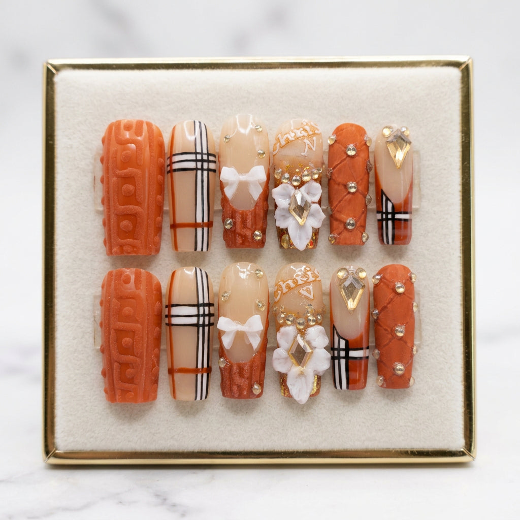 A set of 12 decorative press-on nails displayed in a clear plastic case. The nails feature a mix of orange, beige, and white designs with black-and-white plaid stripes, white bows, 3D floral elements, rhinestones, and gold foil accents.