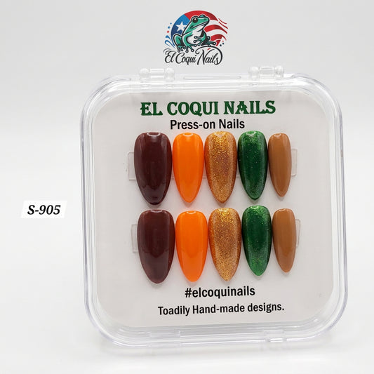 A clear plastic clamshell case of El Coqui Nails press-on nails on a white surface. The nails are arranged in two rows of six and include shades of dark brown, bright orange, metallic gold glitter, emerald green, and tan.