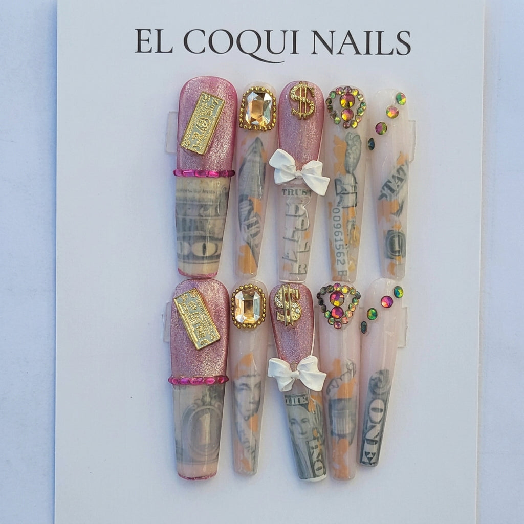 A set of decorative press-on nails displayed on a branded backing card. The nails feature pink glitter, dollar bill print, metallic gold foil, white bows, and colorful rhinestone accents.