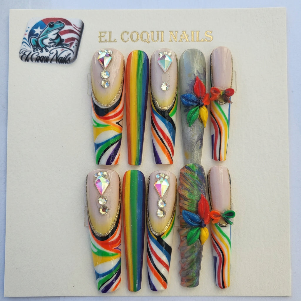 Six decorative press-on nails displayed on a branded card labeled EL COQUINAILS. The nails feature rainbow stripe patterns, rhinestone embellishments, and multicolor floral accents on a light gray background.